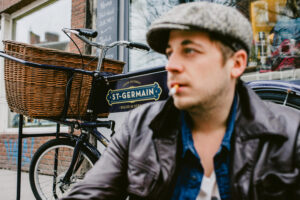Ein Mann mit Zigarette und Schiebermütze sitzt vor einem Restaurant auf Sankt Pauli und hinter ihm an der Wand lehnt ein Vintage Lastenrad mit einem großen Korb am Lenker